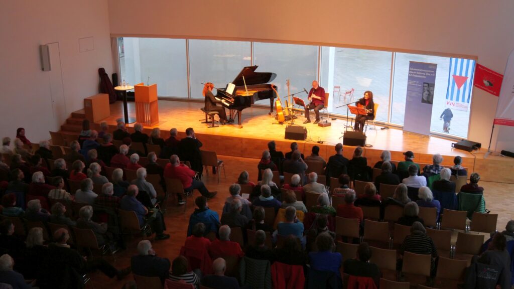 Auf dem Bild ist Turiner Trio „La Desbandá“ bei seinem Auftritt zum 80. Jahrestag der Befreiung vom Faschismus im Stadthaus Ulm zu sehen. Das Trio besteht, wie auf dem Bild zu sehen aus Tiziana Cappellino am Flügel, López Massimo Sartori am Kontrabass und der Gitarristin und Sängerin Angeles Agyado. Das Bild ist eine Totale und zeigt neben dert Bühne auch viele Zuschauerreihen. Foto: Konrad Sedding